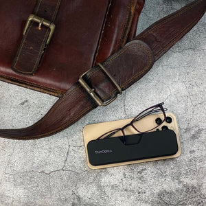 ThinOptics Connect Reading Glasses peeking out of their Connect Case attached to a smartphone on a concrete surface with a leather workbag satchel in frame