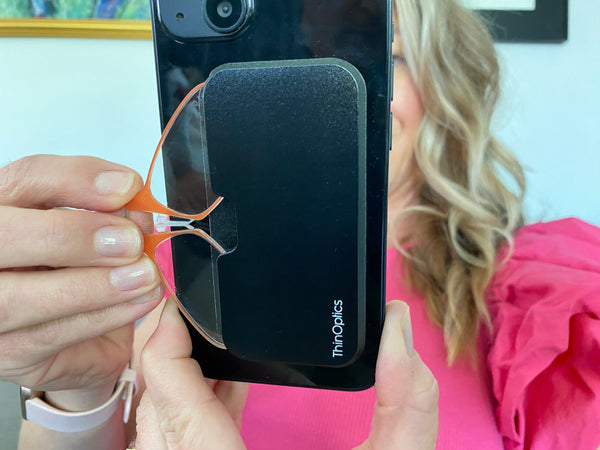 Woman on couch, reading iPhone, retrieving Sunrise Orange Spring Bloom Readers from black Universal Pod on phone - Limited Edition ThinOptics Reading Glasses