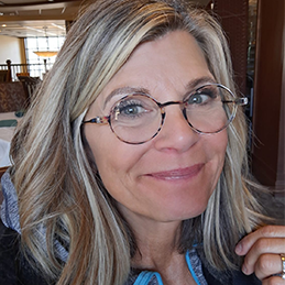 Woman with blonde hair wearing stylish round eyeglasses smiles in restaurant.