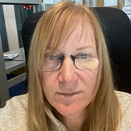 Woman with blonde hair and glasses smiles at the camera from her office chair.