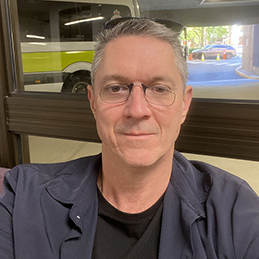 Man with glasses smiles for the camera while sitting indoors. A yellow and white bus is visible through the window behind him.