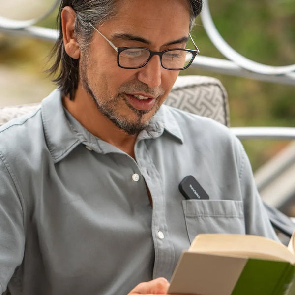 Man wearing glasses enjoys a book outdoors while using a discreet voice amplifier clipped to his shirt.