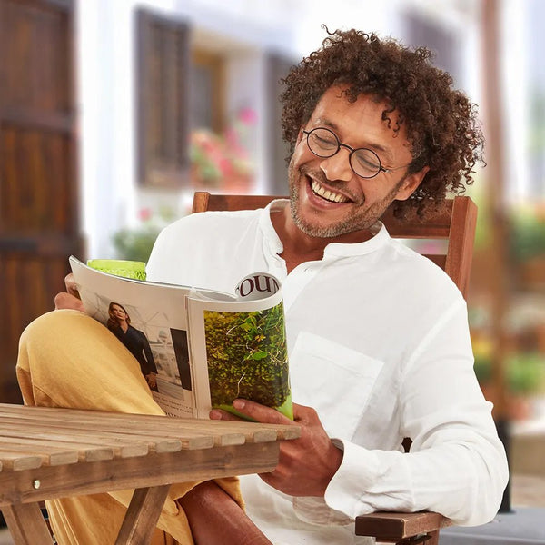 Smiling man with round eyeglasses enjoys reading a magazine outdoors on a patio.