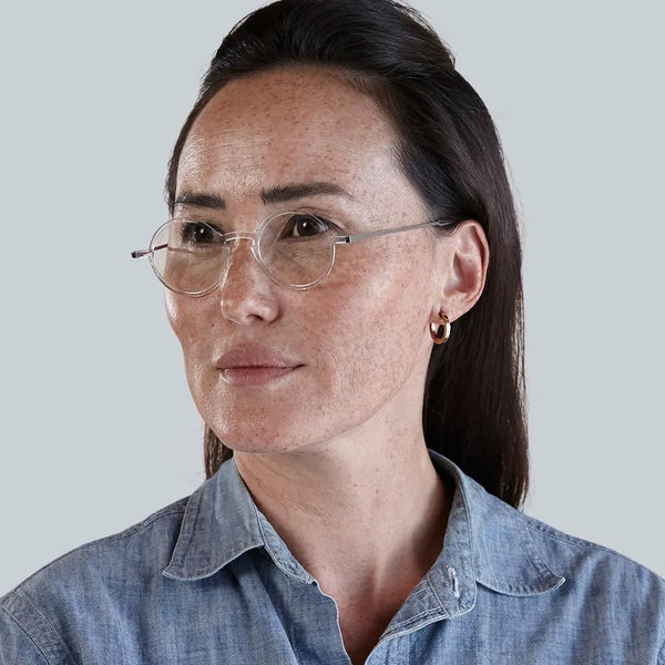 Woman with freckles wears round, clear eyeglasses and a denim shirt.