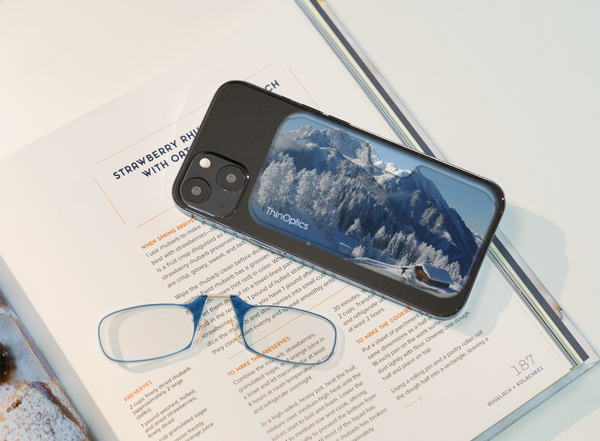 Black iPhone with a snowy mountain scene reflecting on the back glass lies on an open cookbook with a pair of reading glasses.