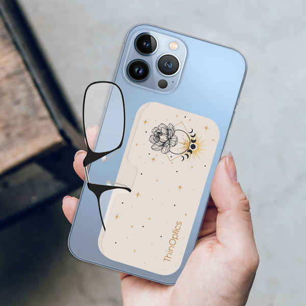 A hand holds a blue smartphone with a ThinOptics case and a pair of reading glasses attached. The case features a celestial design of a flower, moon phases, and stars.