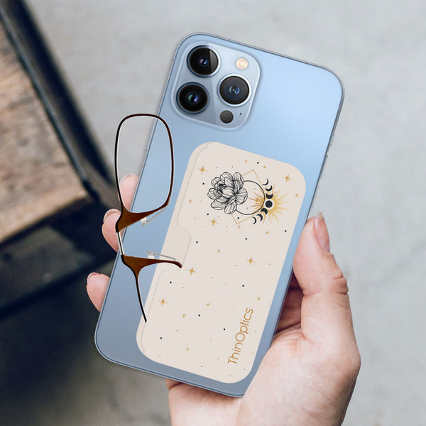 Hand holds blue smartphone with attached ThinOptics reading glasses featuring a celestial floral design.