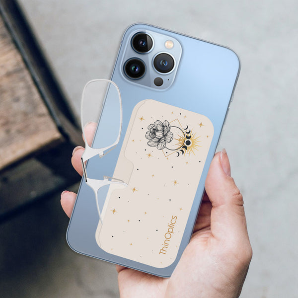 Hand holds a blue smartphone with ThinOptics readers attached to the back. The case features a celestial design with a flower, moon phases, and stars.