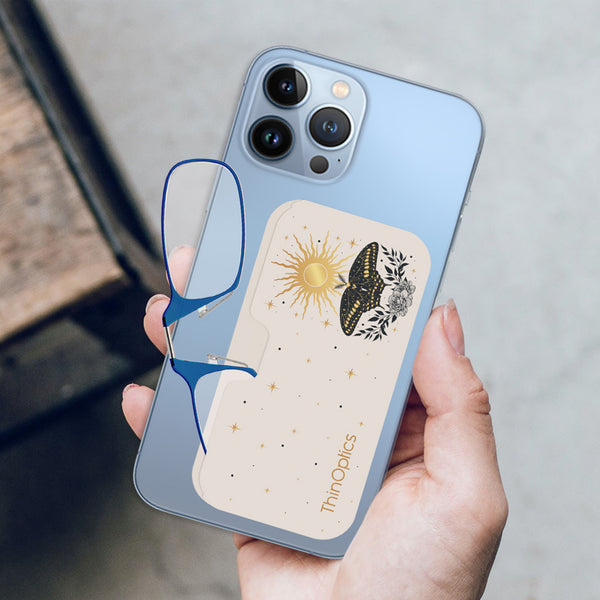 Hand holds a blue smartphone with a ThinOptics reader case featuring a gold sun, butterfly, and floral design attached to the back. Blue ThinOptics readers are tucked into the case.