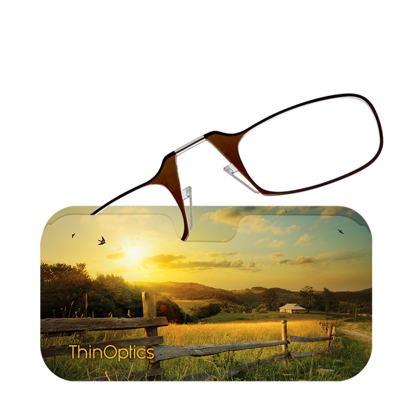 ThinOptics brown reading glasses rest on a serene countryside scene with a golden sunset.
