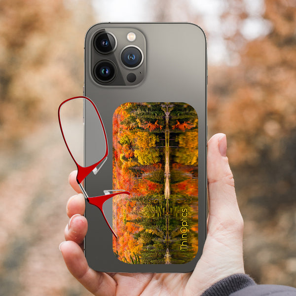 Hand holds a smartphone with a ThinOptics reading glasses case displaying a scenic autumn forest reflected in a lake. Red reading glasses are folded nearby.