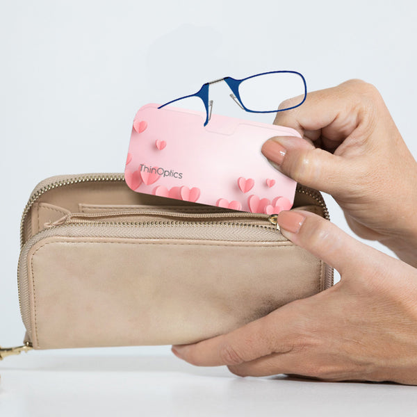 Woman places blue ThinOptics reading glasses into their slim pink heart-patterned case, demonstrating the compact size and portability of the eyewear.