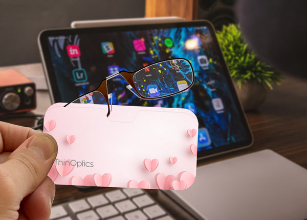 Hand holds ThinOptics reading glasses tucked into sleek, pink heart-patterned case in front of a tablet.