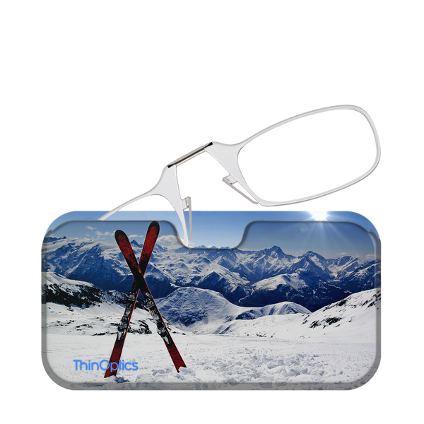 Silver ThinOptics reading glasses folded atop a snowy mountain scene with skis.