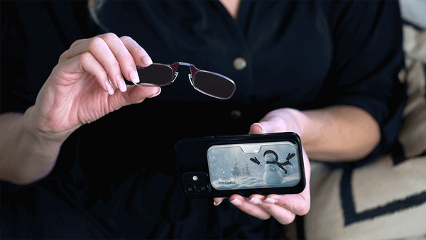 Woman holds ThinOptics reading glasses over a smartphone displaying a winter scene.