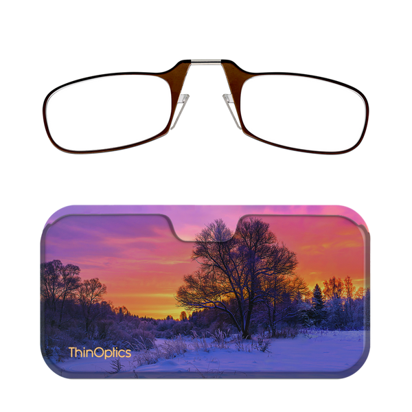 Brown ThinOptics reading glasses rest above a slim case featuring a vibrant sunset over a snow-covered forest.