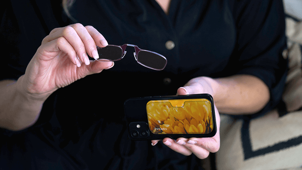 Woman holds ThinOptics reading glasses and a smartphone with a yellow floral ThinOptics case.