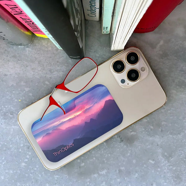 Red ThinOptics reading glasses rest on a smartphone displaying a scenic mountain sunset.