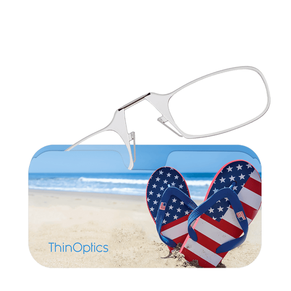 Silver ThinOptics reading glasses rest on a beach-themed case featuring American flag flip-flops nestled in the sand.