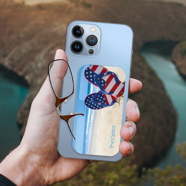 Hand holds a smartphone with a ThinOptics reader attached to the back. The reader features a beach scene with American flag flip flops.