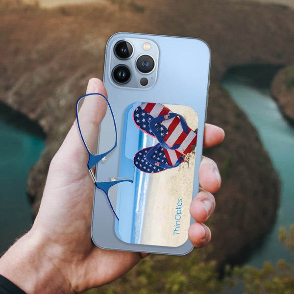 Hand holds a blue smartphone with a ThinOptics case featuring patriotic flip flops on a beach. A pair of ThinOptics reading glasses are tucked into the side.