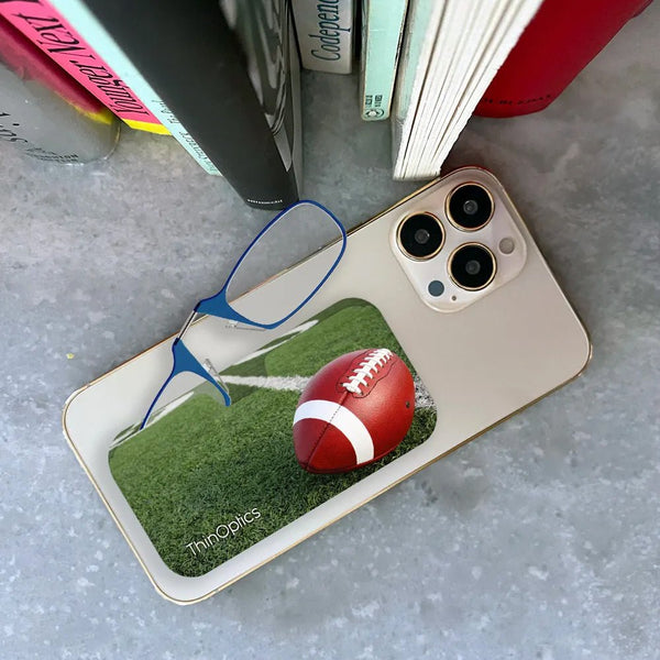 ThinOptics football-themed reading glasses case rests on a smartphone beside blue reading glasses.