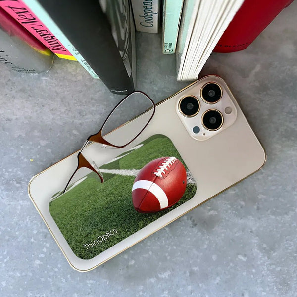 A ThinOptics reading glasses case with a football field design and a football resting on the yard lines sits on top of a smartphone.