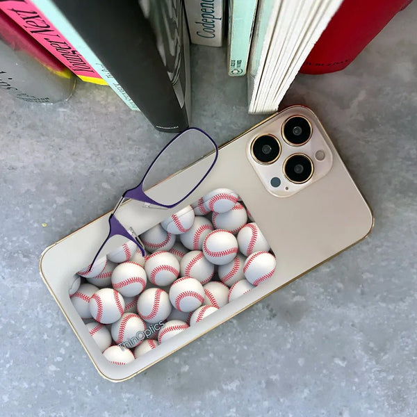 A smartphone with a clear case featuring an optical illusion of baseballs overflowing from the phone's charging port. ThinOptics reading glasses rest on top, highlighting the case's playful design.