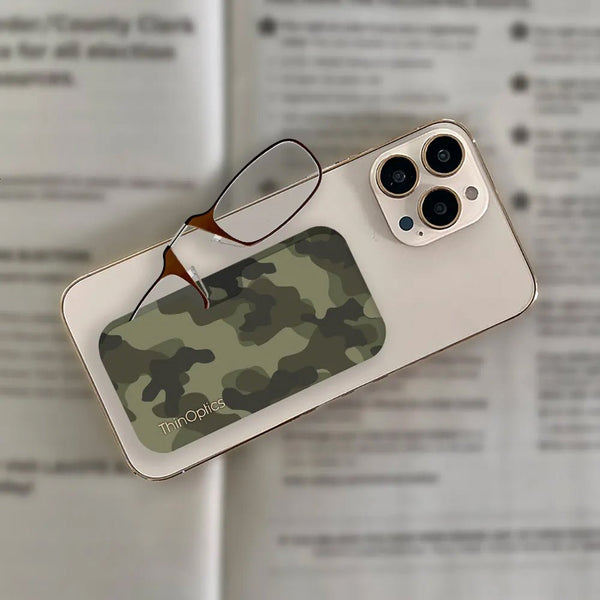 A pair of ThinOptics reading glasses with a camouflage case rests on top of a smartphone on a blurred text background.