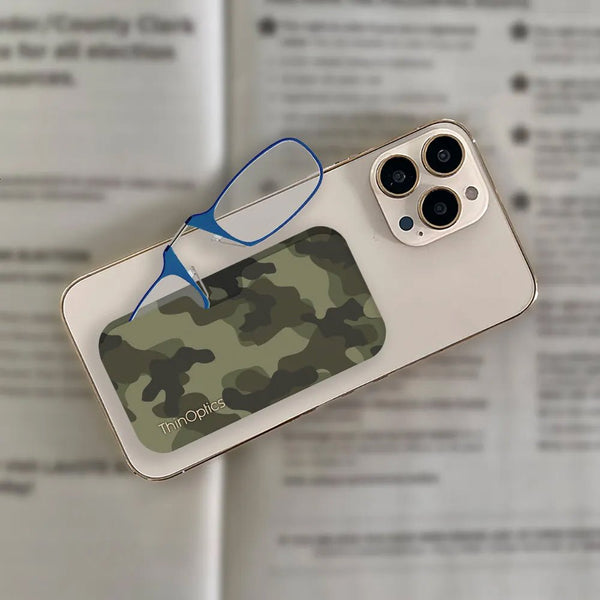 A pair of blue ThinOptics reading glasses rest on a smartphone with a green camouflage case. The glasses are folded and tucked into the adhesive sleeve on the back of the phone case.