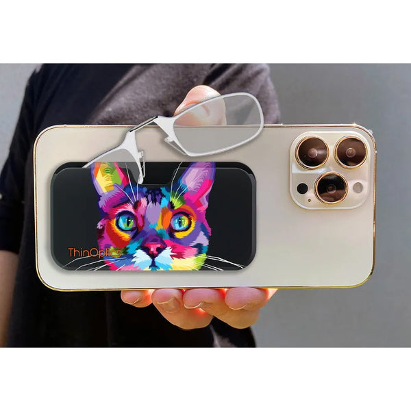Person holds a phone with a ThinOptics reader case featuring a colorful, abstract cat design. Reading glasses rest on top of the case.