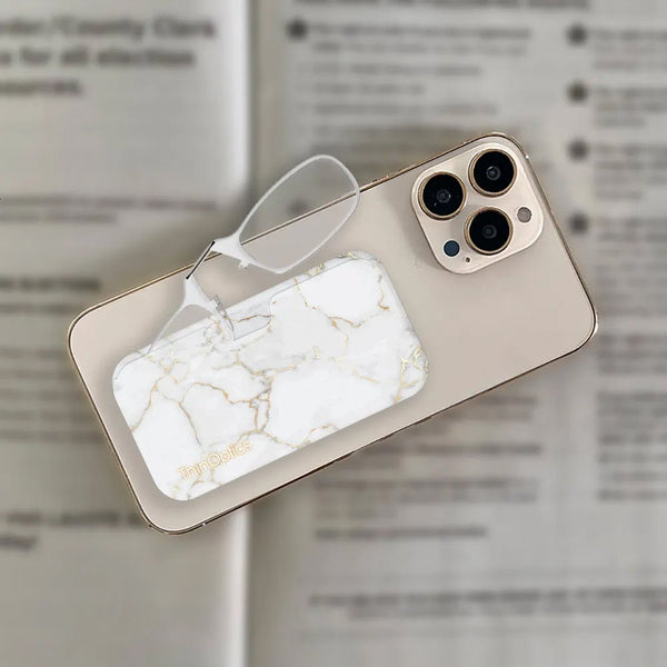 White marble ThinOptics reading glasses case and compact readers rest on a gold smartphone displaying three camera lenses.