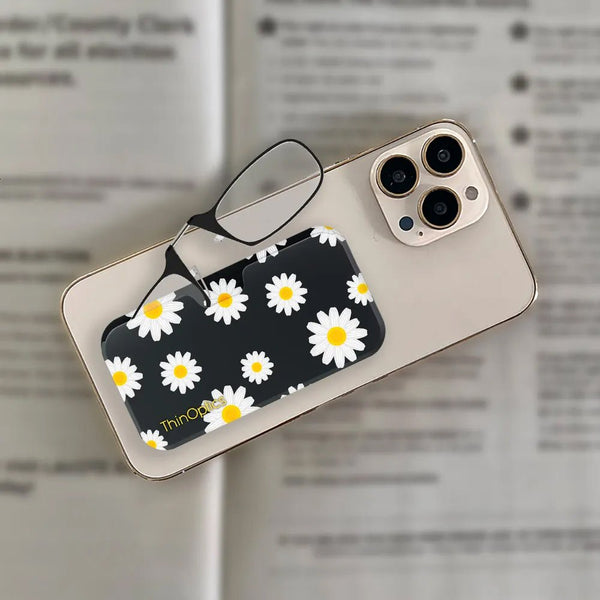 Black and white daisy phone case with attached slim reading glasses resting on open book.