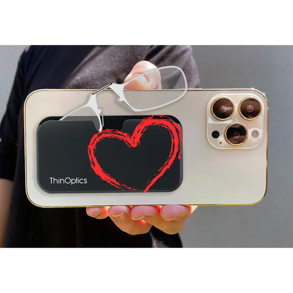 Hand holds a smartphone with a ThinOptics case and a pair of reading glasses. The black case features a red heart design.
