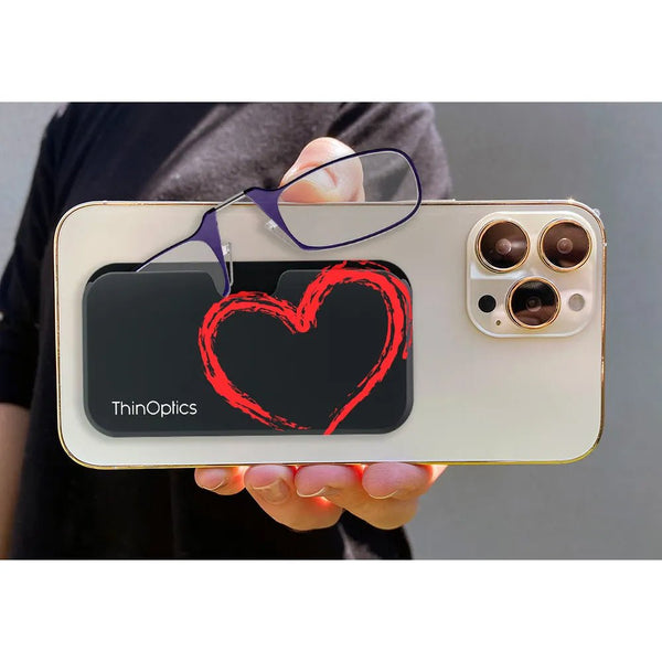 Hand holds smartphone with attached ThinOptics reading glasses and a black case featuring a red heart design.