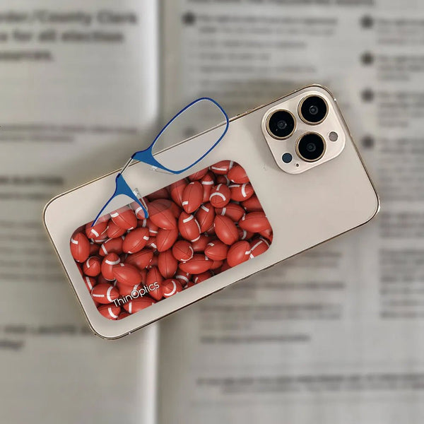 ThinOptics reading glasses rest on a smartphone case filled with miniature footballs, displayed on an open book.