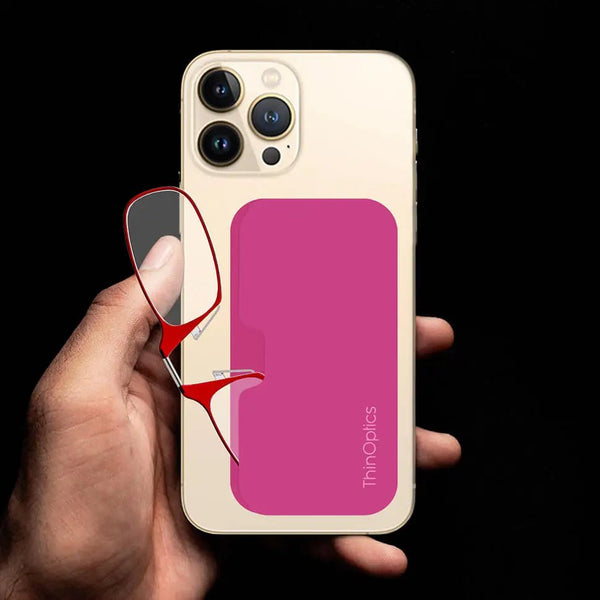 Hand holds a gold smartphone with a ThinOptics pink slimline case attached. Red reading glasses are shown folded and tucked into the case.