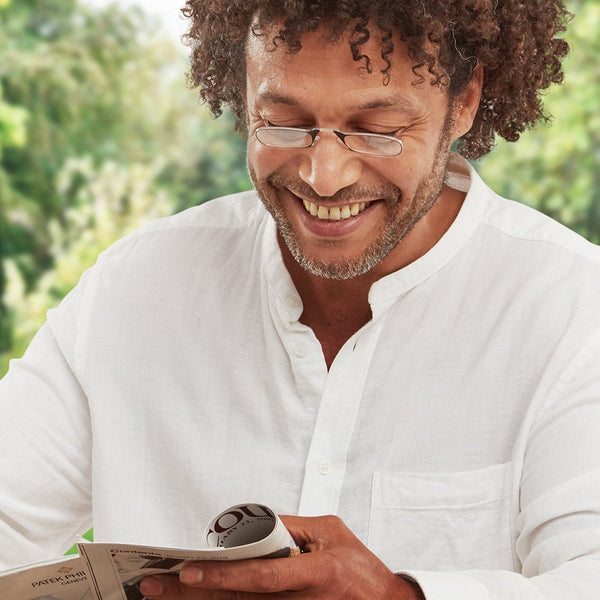 Man with slim reading glasses smiles while enjoying a magazine outdoors.