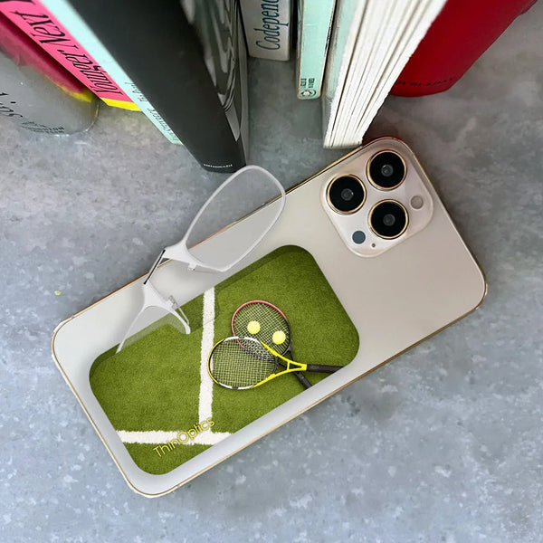 ThinOptics readers rest on a smartphone displaying a tennis court with two rackets and tennis balls.