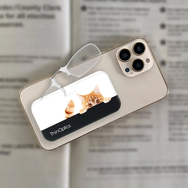 A playful orange tabby cat peeks over a ThinOptics reading glasses case and phone wallet attached to the back of a smartphone.