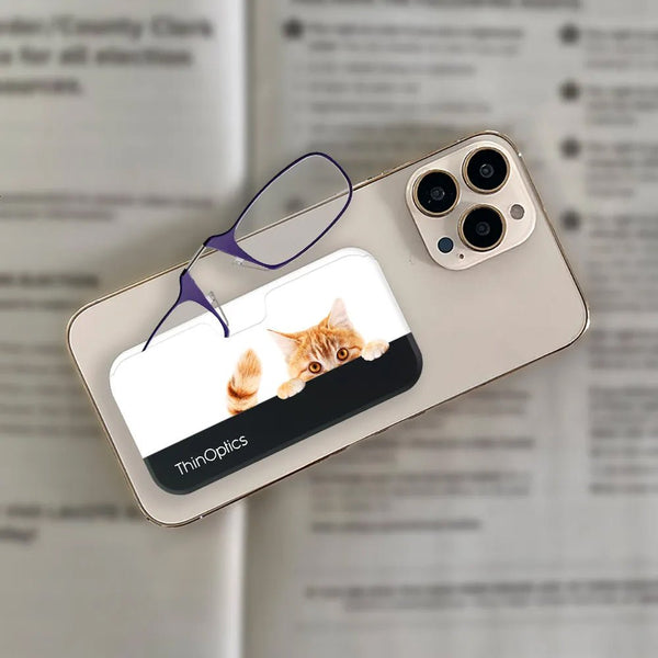 ThinOptics reading glasses with a playful orange tabby cat peeking out from the slim case resting on a smartphone.