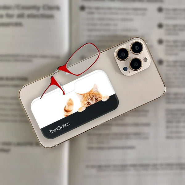 Red ThinOptics reading glasses rest on a smartphone case featuring a playful orange tabby cat peering over the edge.