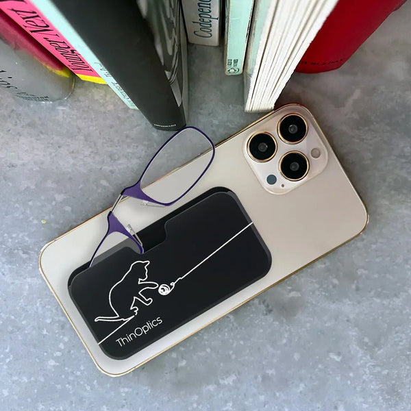ThinOptics reading glasses with purple frames rest on a slim black case featuring a cat playing with yarn. The case sits on a smartphone, ready to slip into a pocket.
