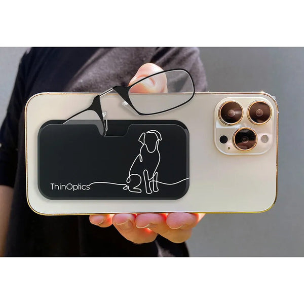 Hand holds smartphone with ThinOptics case and reading glasses attached. The black case features a minimalist line drawing of a dog.