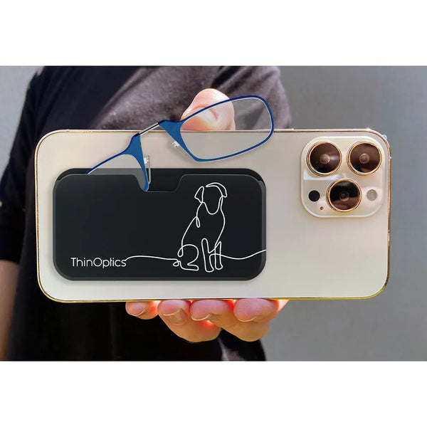 Hand holds a phone with a ThinOptics reading glasses case attached. The case features a minimalist dog design and holds a pair of blue reading glasses.