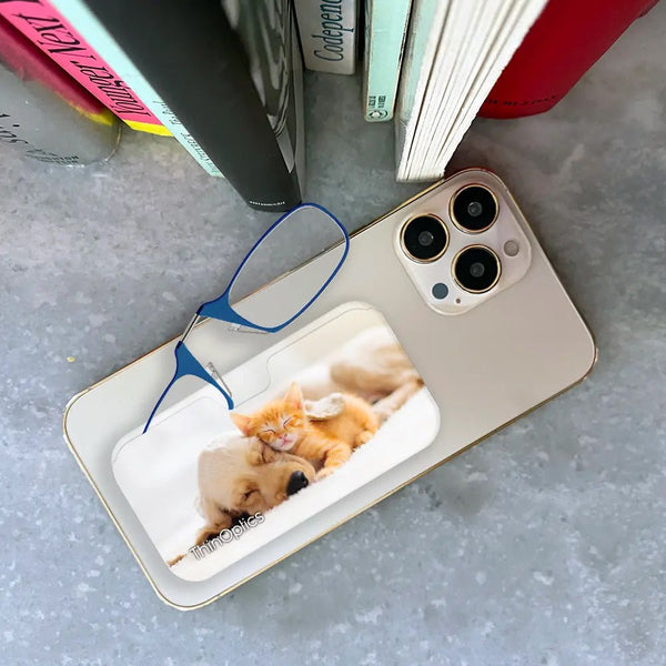 ThinOptics reading glasses rest on a smartphone displaying a heartwarming photo of a sleeping kitten cuddling a puppy.