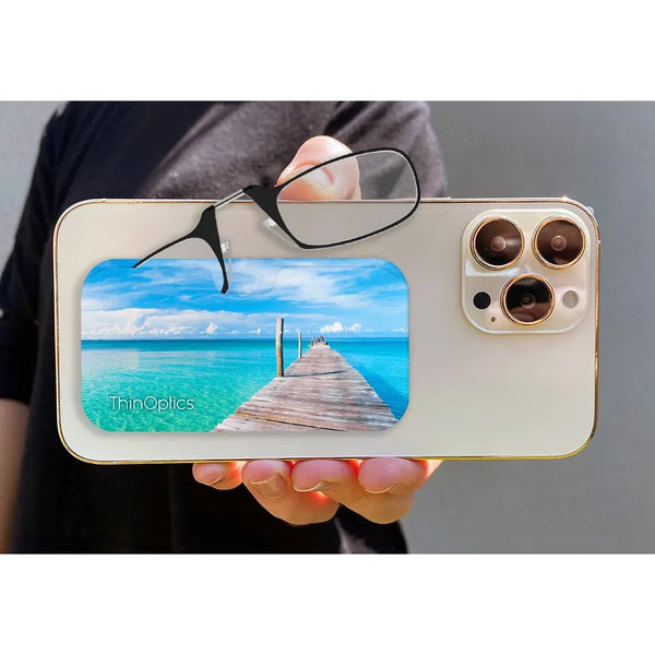 Hand holds ThinOptics readers folded on back of smartphone showcasing vibrant photo of a wooden pier extending over turquoise ocean water.