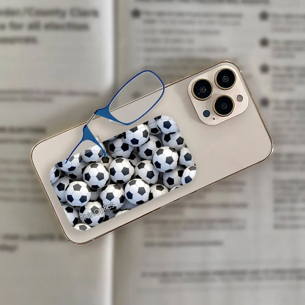 A gold smartphone with a slim, soccer ball-patterned ThinOptics reading glasses case and a pair of blue ThinOptics readers rests on an open book.