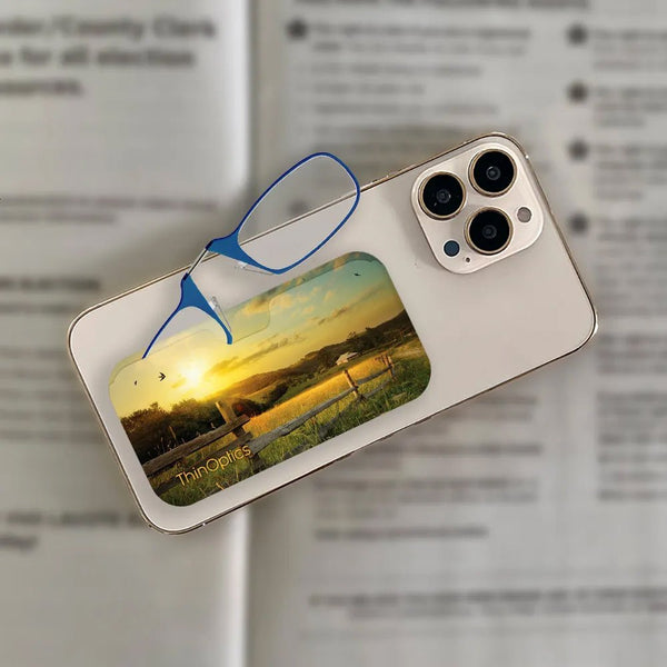 ThinOptics blue reading glasses rest on a smartphone with a sunset farm scene card holder attached.