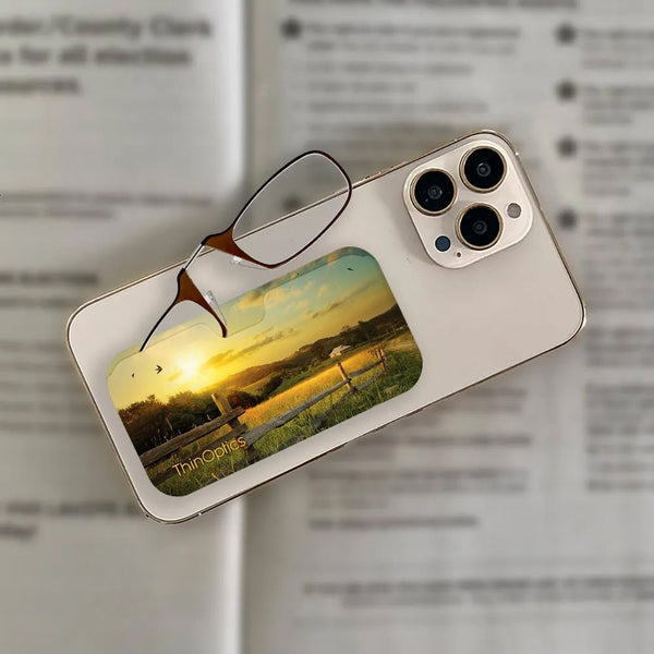 ThinOptics reading glasses resting on a smartphone case displaying a sunset landscape with a farm and fence.